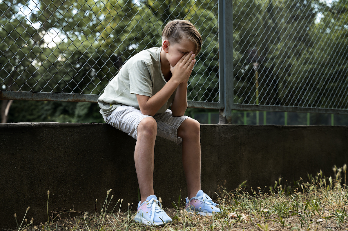 Nen sentat a l'aire lliure amb les mans davant de la cara, amb la mirada cap a terra